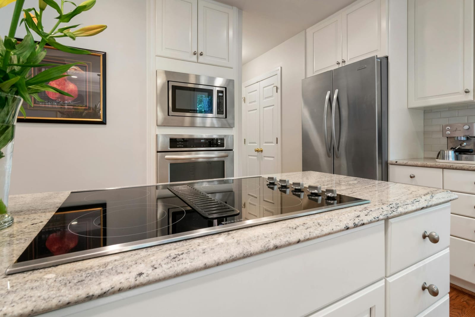 Modern white kitchen with induction cooktop on a granite island, built-in wall oven and microwave, and a stainless side-by-side refrigerator